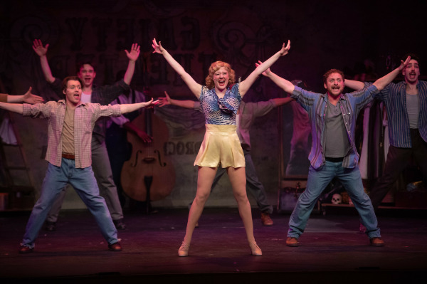 In this image from Crazy For You, a high-energy musical number featuring a smiling performer in the spotlight wearing a blue patterned top and gold shorts, with arms raised in a triumphant pose. Surrounding her, a group of dancers in casual vintage-style costumes mirror her movement, arms extended wide. The background includes stage props like a double bass and a costume rack, suggesting a backstage or rehearsal setting. The lighting and choreography convey excitement and theatrical flair.