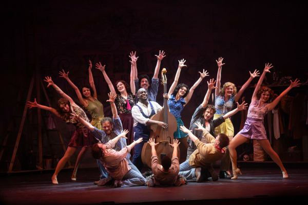 In this image from Crazy For You, a vibrant musical theatre performance featuring a large ensemble cast in a dynamic pose. Centre stage, a performer stands playing an upright bass, surrounded by dancers with wide smiles and arms extended high in a celebratory fan shape. The cast wears colourful, retro-style costumes, and the stage is lit with warm, theatrical lighting against a backdrop suggesting a backstage or rehearsal space, filled with props and costume racks. The image captures the energy and joy of a show-stopping number.