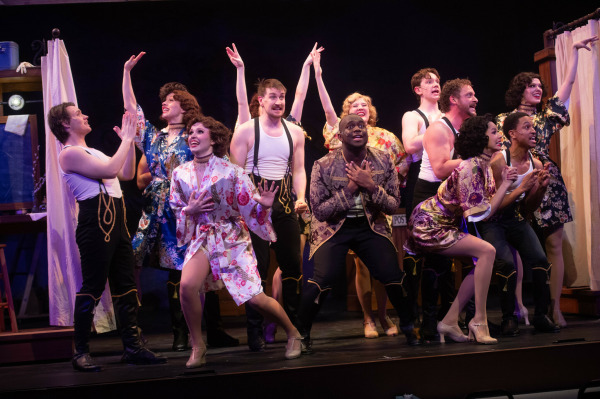 In this image from Crazy For You, A lively group performance on stage featuring a large ensemble cast in theatrical costume. Performers wear floral robes, suspenders, and stylized dancewear, striking expressive poses with wide smiles and animated gestures. The central performer holds their hands to their chest in an emotional moment, while others extend arms upward or gaze outward in joy. The background suggests a backstage dressing room with vanity mirrors and curtains, enhancing the vibrant and playful energy of the scene.