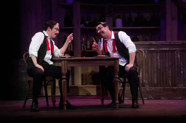 In this image from Crazy For You, a comedic theatre scene featuring two actors dressed identically in white shirts, black vests, black pants, and red neckties, seated at a small wooden table. They are mid-conversation, each pointing a finger while making exaggerated facial expressions. The table holds a bottle, two shot glasses, and plates, set against a rustic bar backdrop filled with bottles and barrels. The lighting and staging suggest a lively and humorous exchange.