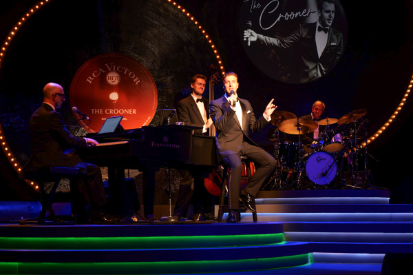 A crooner in a tuxedo sings into a microphone while seated on a stool at centre stage, gesturing with one hand. He’s surrounded by a live band: a pianist on a grand piano, a bassist playing a double bass, and a drummer at a full drum kit. Behind them, a giant vinyl record prop reads “RCA Victor – The Crooner – Michael Vanhevel,” with a second projection of the singer’s image and show title above the drums. The set is illuminated with glowing ring lights and atmospheric stage lighting.