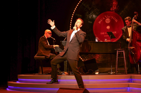 Michael Vanhevel performs centre stage in a dynamic pose, mid-song, with one hand raised dramatically and the other holding a microphone. He wears a classic black tuxedo. Behind him, the pianist plays a grand piano marked 