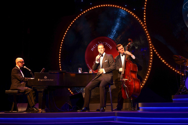 Michael Vanhevel performs in Drayton Entertainment’s The Crooner, seated on a stool beside a grand piano while singing into a microphone. He wears a classic black tuxedo with satin lapels and a white pocket square. To his left, a pianist plays the grand piano with a joyful expression, and a bassist stands behind him, playing an upright bass. The stage backdrop features a large, illuminated vinyl record labelled “RCA Victor – The Crooner,” creating a retro concert atmosphere.
