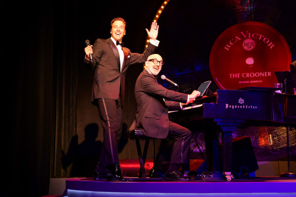 Michael Vanhevel, dressed in a black tuxedo with satin lapels, stands with a microphone in one hand and his other arm raised joyfully while smiling toward the audience. To his left, pianist Mark Payne, also in a black suit, plays a glossy black grand piano while singing into a microphone. Behind them is a giant RCA Victor vinyl record graphic that reads “The Crooner – Michael Vanhevel.” The stage lighting casts a warm red and amber glow, evoking a classic nightclub vibe.