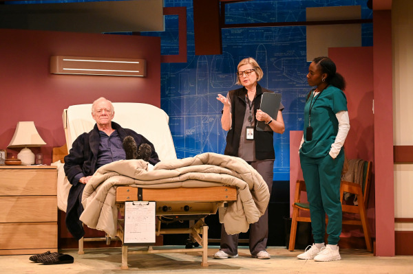  A hospital room scene featuring three characters. An elderly man sits upright in a hospital bed, dressed in dark pyjamas and a robe, with slippers on the floor nearby and a beige comforter covering his legs. Two women stand to his right: one in medical scrubs and the other in a grey uniform holding a clipboard, suggesting a nurse and a doctor or case worker. The setting includes a mauve wall and a backdrop with technical blueprint-style drawings of an aircraft, reinforcing the production’s title and themes. Warm lighting highlights the characters' expressions during a moment of discussion.