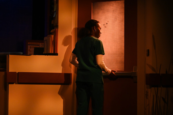 A poignant moment from Flight Risk shows a nurse in green scrubs standing quietly at a closed hospital door, silhouetted by the amber light streaming through the textured glass. Her reflective posture hints at a moment of emotional weight or transition.