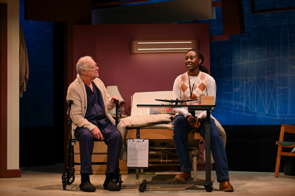 An older man in navy pyjamas and a cardigan sits on a mobility walker in a hospital room, smiling at a young woman seated on his hospital bed. She wears an argyle vest and jeans, and sits beside a model airplane and a large book on an overbed table. The walls behind them are decorated with blueprints of aircraft, and the mood is warm and intimate under soft lighting.