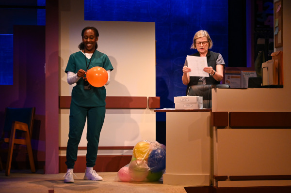 A scene from Flight Risk, featuring an older man in pyjamas and a cardigan seated on a walker next to a hospital bed, smiling at a young nurse or caregiver sitting on the edge of the bed. The nurse wears an argyle sweater vest and has a model airplane and a stack of books on the tray table. The backdrop includes blueprints of aircraft parts, reinforcing the aviation theme of the show. Warm lighting and set details evoke a hospital room setting.