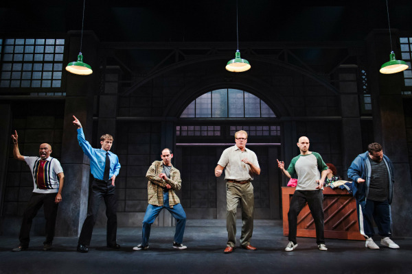  A comedic and energetic stage scene featuring six performers in casual, mismatched outfits, mid-dance with exaggerated poses. The setting resembles a gritty industrial space with large factory windows, concrete columns, and hanging green lights. Each performer strikes a unique pose, evoking confidence, hesitation, or excitement, capturing the humorous spirit of a rehearsal or transformation moment from The Full Monty. A piano and costume rack are visible in the background.