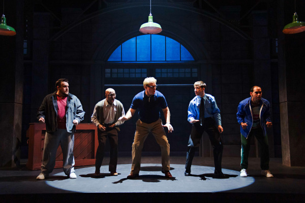 A dramatic and humorous stage moment from The Full Monty. Five performers stand in a row under a spotlight, crouched in intense and determined poses as if preparing for a dance or action sequence. Each is dressed in casual or mismatched clothing, reflecting their diverse characters. The industrial-style set, with large arched windows and hanging green lamps, creates a gritty rehearsal hall atmosphere filled with anticipation and camaraderie.