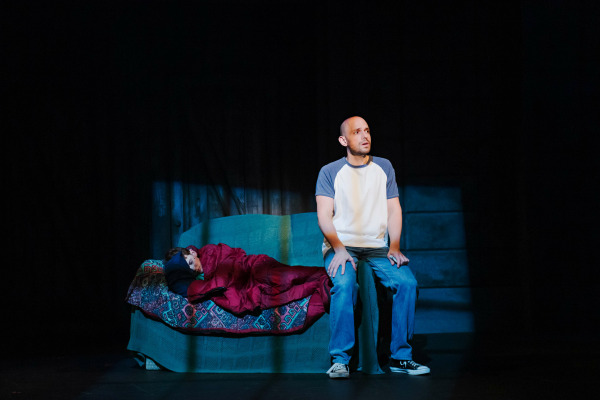 A quiet, emotional theatre moment from The Full Monty. A man in a white and blue ringer t-shirt and jeans sits on the edge of a couch, gazing upward with a pensive expression. Beside him, a child sleeps curled under a red blanket on a patterned quilt. The stage is dimly lit with a spotlight highlighting the man, emphasizing a sense of reflection and tenderness.