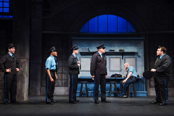 A comedic theatre scene set in a police locker room. Six performers dressed in police uniforms are engaged in animated conversation. Four officers stand in a row facing a man on the far right, who holds a round black hat and appears to be speaking assertively. Another officer sits in the background, deep in thought with his hand on his chin. The set features grey lockers, blue stools, and a large arched window, capturing the look of a precinct interior.