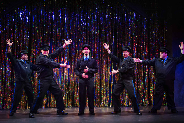 A comedic and theatrical moment from The Full Monty featuring five performers dressed as police officers posing dramatically in front of a shimmering, rainbow-coloured tinsel curtain. The central performer stands still with a serious expression while the others gesture flamboyantly toward him, striking exaggerated stage poses. The sparkling backdrop and playful staging suggest a humorous performance or talent show scene.