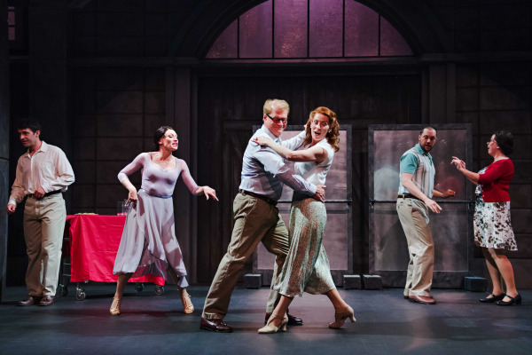  A lighthearted moment from The Full Monty, featuring six performers on stage in mid-action. Centre stage, a couple dances joyfully, with the man in a light blue shirt and the woman in a floral skirt and heels. To the left, a woman in a lavender dress gestures expressively while another man looks on near a red-covered table. On the right, another couple interacts with surprised expressions near metal double doors. The set has an industrial look, with a large arched window glowing with purple light in the background, suggesting a transformed factory or community hall setting.