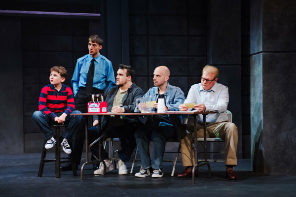  A stage scene from The Full Monty showing five men and a young boy gathered around a folding table. The boy, in a red and navy striped shirt, sits at one end, while the others—dressed in casual clothes and one in a police uniform—sit or stand with attentive or concerned expressions. The table holds a six-pack of beer, a water pitcher, paper cups, and a bowl. The setting suggests a serious or reflective group discussion in a modest, industrial environment.