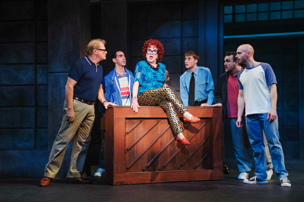 A comedic stage moment from The Full Monty featuring a lively group scene. A character in a bold blue top, patterned gold pants, and red heels with curly red hair and large glasses sits confidently atop a wooden piano. Six men stand around her in various casual outfits, reacting with animated expressions of surprise, curiosity, and disbelief. The industrial-style set and dramatic lighting highlight the humorous and energetic tone of the scene.