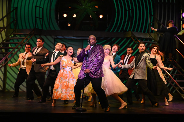 A vibrant scene from Grease captures the high-energy excitement of the school dance. Centre stage, a performer in a sparkly purple blazer sings into a vintage microphone as a lively ensemble of students in colourful retro formalwear dance around him. The backdrop features bold green and black swirls, with palm trees silhouetted behind the raised platform. The joyful expressions and dynamic poses reflect the fun and flirtation of a 1950s prom night brought to life on stage.