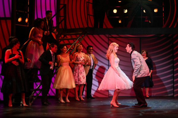 A joyful moment from Grease captures a key dance scene as Sandy and Danny face each other, smiling and mid-step, dressed in classic 1950s prom attire. Sandy wears a sparkling pink party dress, while Danny sports a silver-striped blazer. Around them, fellow students in formal dresses and suits cheer them on against a bold red and purple striped backdrop, enhancing the excitement and energy of the school dance setting.