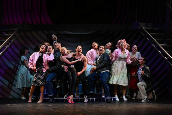 A high-energy moment from Grease shows the full ensemble gathered around centre stage, beaming with joy during the show’s grand finale. Sandy appears in her iconic black outfit with red heels, sitting on a bench alongside Danny, who wears a black tank top and jeans. Surrounding them are the Pink Ladies in their pink jackets and the T-Birds in leather, all caught mid-song with arms around each other in a pose full of celebration and camaraderie. The vivid lighting and diner-inspired set complete the electrifying scene.