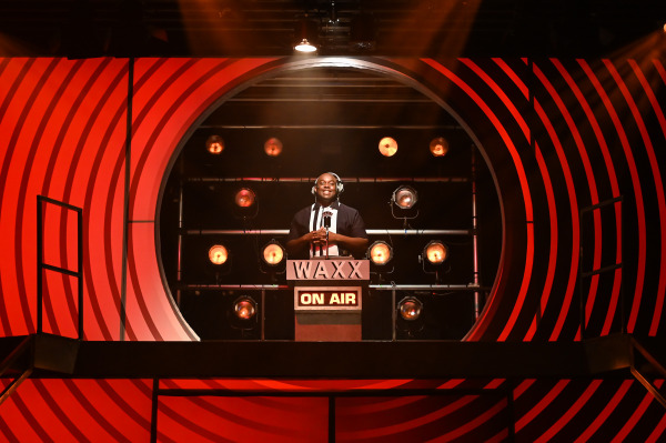 A performance scene from Grease features a smiling DJ standing at a vintage radio booth marked “WAXX” and “ON AIR.” Wearing headphones and a black and white outfit, the performer stands confidently under a spotlight, framed by concentric red stripes and surrounded by glowing stage lights. The dynamic, retro-inspired set evokes the energy of a live radio broadcast, setting the tone for the show's rock 'n' roll era.