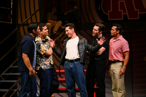 A high-energy performance scene from Grease features five male performers gathered mid-conversation on stage. At the center, a performer in a white T-shirt, jeans, and a black leather jacket gestures animatedly with open hands, exuding confidence. The others surround him in casual 1950s-style attire—one in a Hawaiian shirt, one in a dark bowling shirt, and two in solid black and red shirts—each reacting with amusement or curiosity. The retro set features red stairs and a striped backdrop, evoking the lively atmosphere of Rydell High.