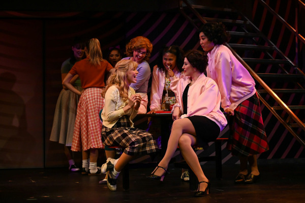 A vibrant scene from Grease shows eight performers gathered around a diner table mid-conversation. At the centre, one performer in a yellow cardigan and plaid skirt sits animatedly chatting with another seated performer in a black dress and pink satin jacket. The surrounding ensemble, also dressed in classic 1950s outfits with full skirts and Pink Ladies jackets, lean in eagerly, smiling and sipping from a Coca-Cola bottle. The backdrop features retro stripes and a metal staircase, capturing the youthful energy and camaraderie of the iconic musical.