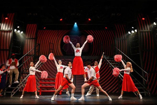    A high-energy scene from Grease features a cheerleading routine in full swing, with a performer lifted high in the centre holding red and white pom-poms. Surrounding her, cheerleaders in matching red skirts and “R” sweaters strike coordinated poses, while two male students in white athletic wear anchor the lift. The stylized red-and-black striped set and bold school spirit create a lively, vintage high school atmosphere.