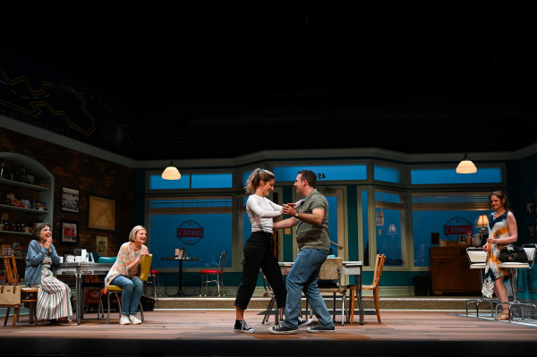 A moment of joy unfolds in a warmly lit diner as two characters dance playfully at centre stage, holding hands and smiling at each other. Three others watch from opposite sides of the stage—two seated women laugh and clap in delight on the left, while another character in a colourful dress looks on fondly from the right. The set features retro diner furnishings, teal walls, large windows, and a welcoming atmosphere.