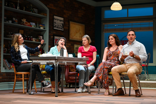  Five characters are seated around a table in a cozy diner setting. One person dramatically rolls their eyes with a comical expression while the others react with amusement or surprise. The backdrop includes brick walls, framed photographs, and shelves filled with preserves and books, creating a warm, lived-in atmosphere. A slice of pie is held mid-bite by the person on the far right, adding to the lighthearted tone of the scene.