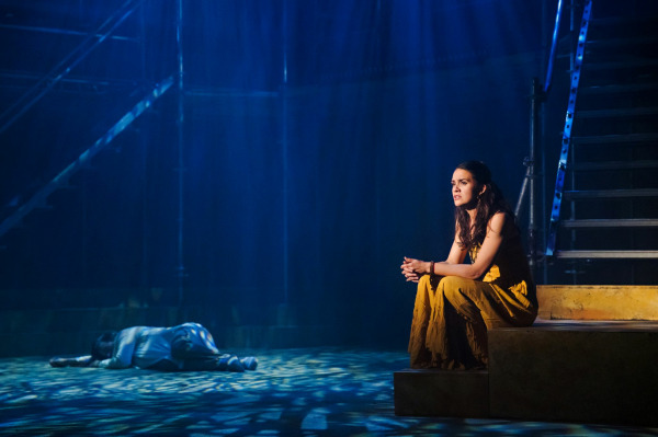 A poignant scene from Jesus Christ Superstar featuring a performer in a flowing mustard yellow dress sitting on a step, looking out in contemplation under soft, moody blue lighting. In the background, another performer lies face down on the ground, partially illuminated. The industrial set, with metal scaffolding and stairs, evokes a modern, minimalist atmosphere.