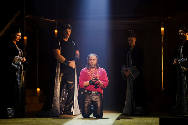 A powerful theatrical moment from Jesus Christ Superstar shows a performer kneeling center stage in a spotlight, wearing a vivid red shirt with gold trim and black leather pants. Their expression is intense and emotional, with hands clasped as if in plea or reflection. Surrounding them are five figures dressed in black robes with silver trim and crowns, standing solemnly in the shadows. The dimly lit set is accented by glowing candlelight, enhancing the dramatic, reverent atmosphere.