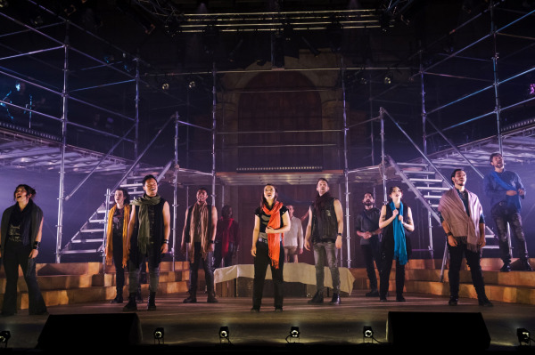   A wide ensemble moment from Jesus Christ Superstar, featuring a group of performers standing in a powerful formation on a dimly lit stage framed by industrial scaffolding. Each cast member wears modernized garments in earthy tones with colourful scarves, and they appear mid-song, conveying emotional intensity. The lighting casts a soft haze across the set, highlighting the contemporary and dramatic aesthetic of the production.