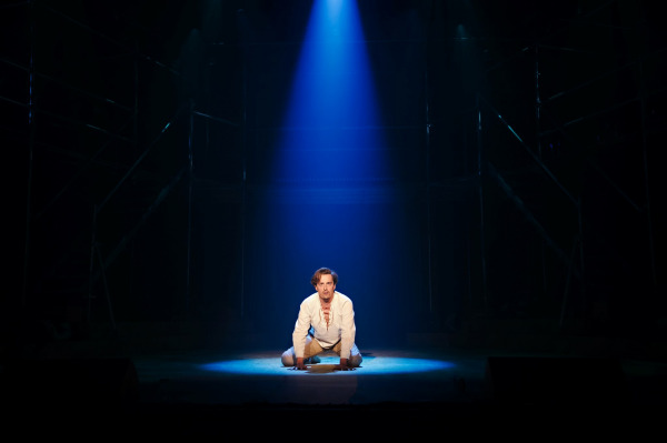 A powerful solo moment from Jesus Christ Superstar, featuring a performer in a white tunic with lace-up neckline kneeling center stage under a dramatic blue spotlight. The surrounding stage is dark, with industrial scaffolding faintly visible in the background, emphasizing the emotional intensity and isolation of the scene.
