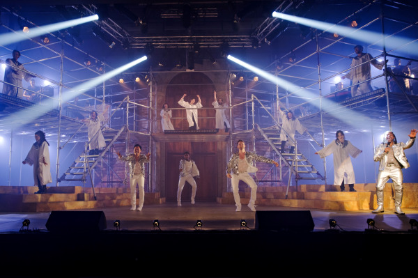 A vibrant ensemble number from Jesus Christ Superstar, with performers spread across a multi-level scaffolding set bathed in bright white and blue stage lights. The cast wears glistening white and silver costumes, with some in sequin jackets and others in long robes. The central platform features dancers in energetic poses, while musicians can be seen in the upper corners. One performer on the far right, wearing a shimmering silver outfit, passionately sings into a microphone. The overall scene radiates a celebratory and electrifying concert-style atmosphere.