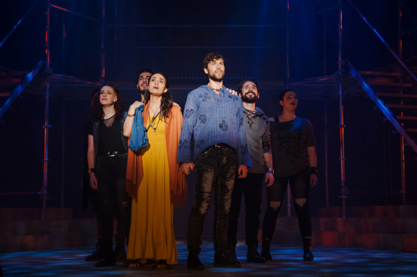 A dramatic stage scene from Jesus Christ Superstar featuring six performers standing close together, illuminated by moody blue and orange lighting. The central figures—a person in a yellow dress with a burnt orange shawl and another in a blue embroidered tunic—stand hand in hand, gazing upward with solemn expressions. The ensemble surrounding them wears contemporary, distressed costumes with dark tones. Behind them, a minimalist scaffolding set suggests an industrial or urban environment.
