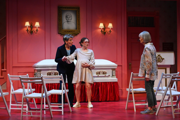 A scene from Liars at a Funeral shows three performers in a confrontation at the front of a funeral parlour. One character in a dark suit holds the hand of a visibly pregnant character in a cream dress and red glasses, who clutches her belly while looking toward an older character in a floral blouse and green pants. White folding chairs are scattered across the red floor, and a white casket with red skirting sits beneath a portrait of the deceased. The pink walls and warm lighting create a contrast between the solemn setting and the heightened tension of the moment.