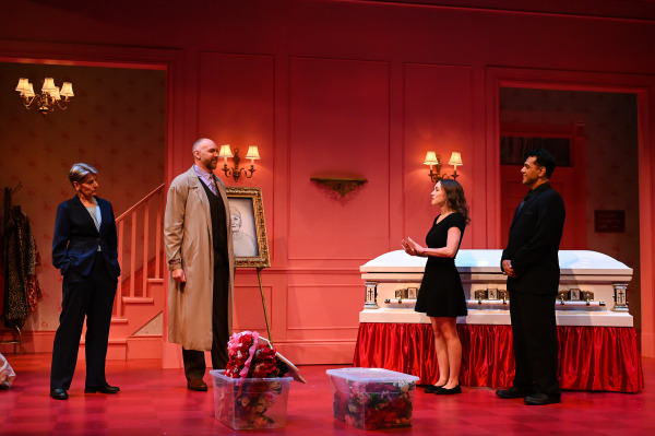A scene from Liars at a Funeral shows four performers in a lavishly decorated funeral parlour with salmon-pink walls, warm wall sconces, and a white casket trimmed in red fabric. Two large bins of artificial flowers sit at the performers’ feet. One performer in a black dress gestures while speaking to a man in a trench coat and another dressed in black. A fourth performer, in a navy pantsuit, watches the exchange near a staircase. A portrait of the deceased is displayed prominently between the wall sconces, adding to the comedic tension of the moment.