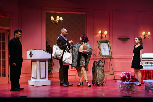 A scene from Liars at a Funeral captures five performers in a vividly pink funeral parlour set. At centre stage, two characters greet each other warmly—one holds multiple tote bags while the other, dressed in animal print and heels, carries a paper bag and stands beside matching zebra-print luggage. A third performer, in a black dress, reacts with surprise, while a fourth stands silently near a white podium. A framed portrait of the deceased and bins of artificial flowers near the casket add to the chaotic, comedic atmosphere.