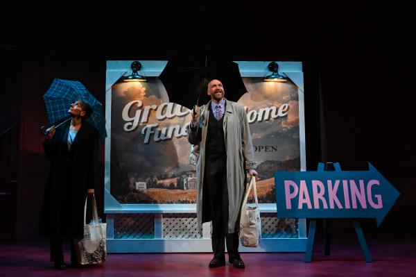 A scene from Liars at a Funeral features two performers standing in front of a painted funeral home sign that reads “Grace Funeral Home.” One holds a black umbrella and smiles brightly, dressed in a long trench coat with a patterned tie and carrying a canvas tote. The other, in a dark coat and slacks, holds a plaid umbrella and looks upward with a curious expression. A large blue arrow-shaped sign marked “PARKING” sits to the right. The backdrop evokes a cloudy sky and rolling hills, with warm lighting suggesting an overcast or rainy day.