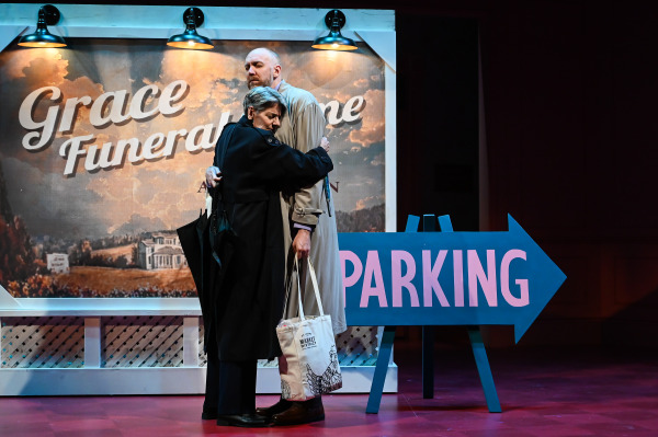 A scene from Liars at a Funeral shows two performers embracing in front of a painted sign that reads “Grace Funeral Home.” The figure on the left wears a dark overcoat and holds a closed umbrella, their expression somber as they lean into the taller figure on the right, who is dressed in a light trench coat and carries a tote bag. A large blue arrow-shaped “PARKING” sign points to the right, and the warm lighting highlights the emotional weight of the moment against a backdrop of rolling hills and cloudy skies.