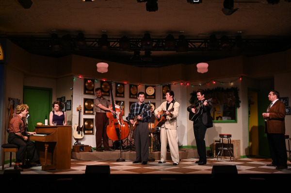   A wide shot from Million Dollar Quartet captures a full ensemble mid-performance in a 1950s recording studio set. One performer plays the piano on the left, with another seated nearby at a barstool. At centre stage, three performers stand with guitars and a mandolin, harmonizing around a vintage microphone. A fourth musician stands behind them playing upright bass, while a fifth plays guitar to the right. A man in a plaid jacket looks on from the far right. The stage is decorated with gold records, Christmas lights, and vintage studio decor, evoking a warm, nostalgic mood.