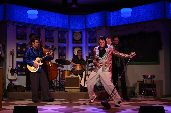 A dynamic performance moment from Million Dollar Quartet shows a performer in a sparkling silver jacket and white pants striking an Elvis-style pose, gripping a tilted microphone stand and dancing across the stage. A guitarist in a glittery blue jacket plays on the left, while a bassist and drummer support from the back. Another cast member stands behind, mid-applause. The set is decorated with gold records and festive holiday lights, enhancing the vintage recording studio atmosphere.