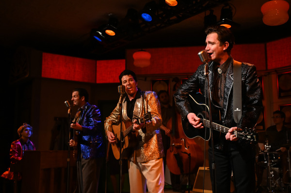 A lively scene from Million Dollar Quartet features four performers at the front of the stage singing into vintage microphones while playing guitars. They are dressed in sparkling jackets—gold, blue, and black—evoking a classic rock-and-roll style. A young performer at the piano and other musicians, including a bassist and drummer, support in the background. The setting resembles a retro recording studio with red accent lighting and a wall lined with gold records. The mood is energetic and celebratory.