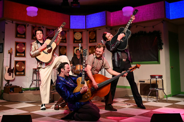 A high-energy performance scene from Million Dollar Quartet features five performers mid-song in dynamic poses. Three guitarists, dressed in glittery jackets, strike dramatic stances while playing their instruments. One kneels on the floor in a blue sequin jacket, another stands with a gold acoustic guitar and a foot propped up on a stool, and the third holds a black guitar overhead. A fourth performer plays an upright bass, leaning in toward the action, while a drummer in the background keeps the rhythm. The stage is styled like a retro recording studio, with gold records lining the wall and warm, colourful lighting.