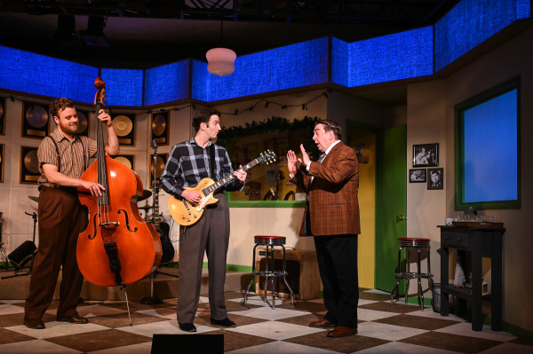 A scene from Million Dollar Quartet set in a retro recording studio. On the left, a musician plays an upright bass with a relaxed expression. At centre, a guitarist in a plaid shirt looks to his right, mid-song or conversation. To the right, a man in a brown plaid jacket gestures animatedly with raised hands, as if giving direction or reacting to the music. The background features gold records on the wall, holiday garland, and framed black-and-white photos. The set has a vintage aesthetic with checkered floors and colourful accents
