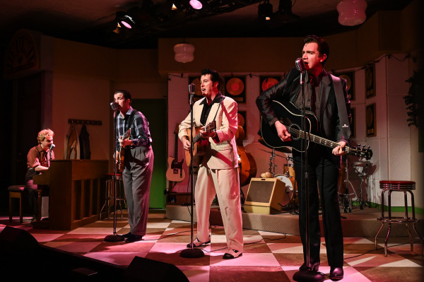 A lively performance scene from Million Dollar Quartet, featuring four musicians at the front of the stage. Three stand with guitars—dressed in period-inspired suits and singing into vintage microphones—while the fourth plays piano to the left. The backdrop resembles a recording studio with guitars hanging on the wall and gold records displayed. Warm stage lighting and checkered flooring enhance the nostalgic 1950s ambiance.