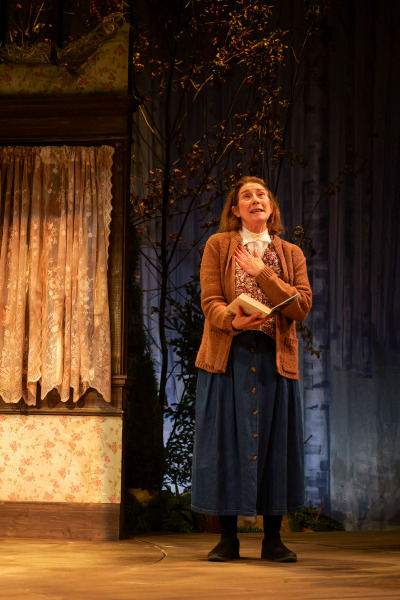 Misery: A woman stands on stage clutching a small book to her chest, gazing upward with an expression of emotion and longing. She wears a brown knit cardigan over a floral blouse and blue buttoned skirt. Behind her, a window with vintage lace curtains and weathered floral wallpaper adds a rustic charm, while barren trees silhouetted in blue light evoke a haunting, isolated atmosphere.