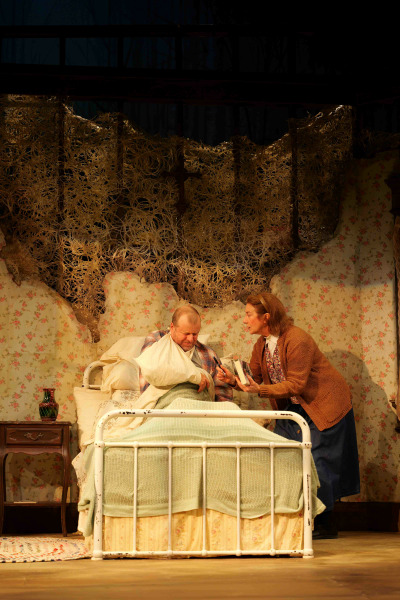 A tense scene from Misery features a bedridden man with a bandaged arm sitting upright in a white metal-framed bed, while a woman leans over him, holding a book and pen as he prepares to write. The floral wallpaper behind them is aged and torn, partially covered by a tangled mesh, creating a sense of claustrophobia and unease. The woman’s cardigan and long denim skirt contrast with the man's plaid shirt and green blanket, hinting at their contrasting roles. The lighting casts warm tones over the disturbing domestic setting, underscoring the psychological intensity of the moment.
