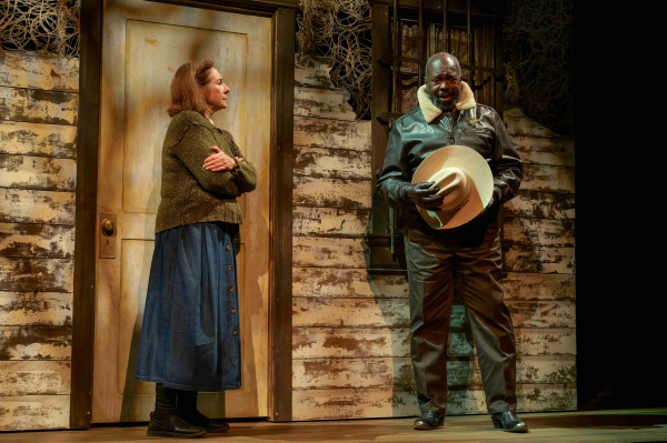 A tense exchange unfolds between Annie Wilkes and Sheriff Buster outside a rustic, weather-worn house. Annie stands rigid with her arms crossed, while the Sheriff, dressed in a leather jacket and holding his wide-brimmed hat, cautiously questions her. The peeling paint on the door and siding, coupled with the dim lighting, heightens the sense of unease and suspense in this pivotal scene.