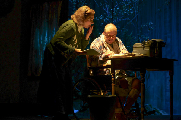 A tense scene from Misery shows a woman looming over a man in a wheelchair at a wooden writing desk. The woman leans in closely, holding a piece of paper with an intense expression as the man, wrapped in a plaid shirt and bandages, focuses on the manuscript in front of him. A vintage typewriter, a glass of water, and a stack of pages complete the ominous setup under a blue-toned light, suggesting both physical confinement and psychological pressure.